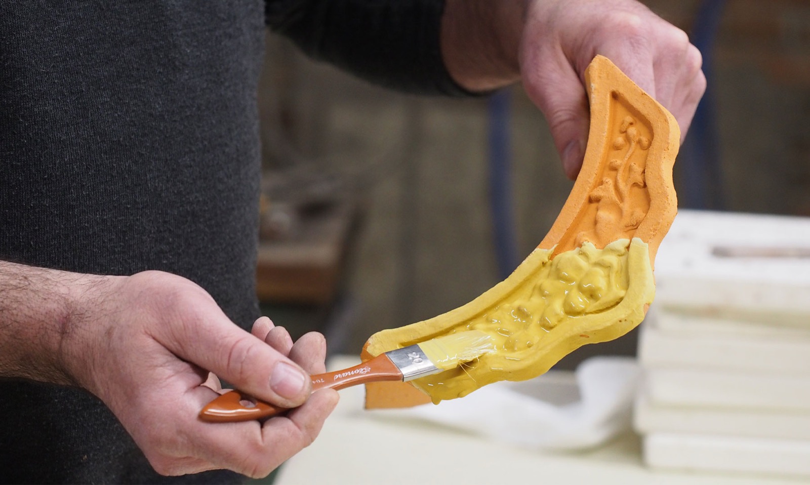 Mains de l'artisan émaillant au pinceau une pièce de céramique architecturale crue.